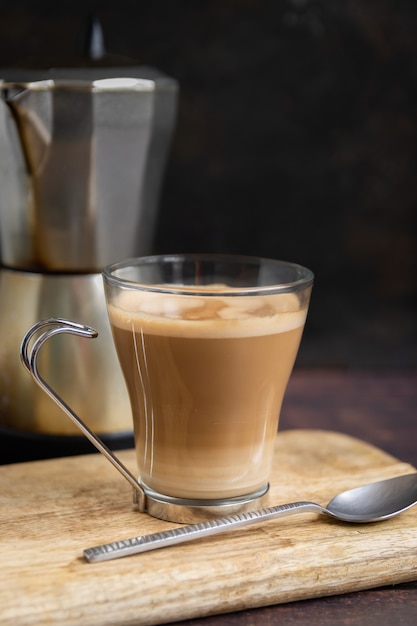 Cup of coffee with milk and teaspoon on wooden table and Italian coffee pot in the background. Vertical