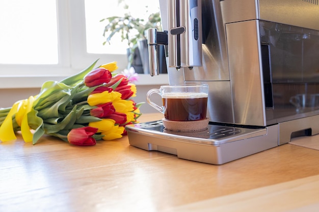 Cup of coffee with milk and tulips flowers on wooden kitchen table Freshly brewed cappuccino espresso in coffee machine for breakfast Stylish home interior decor