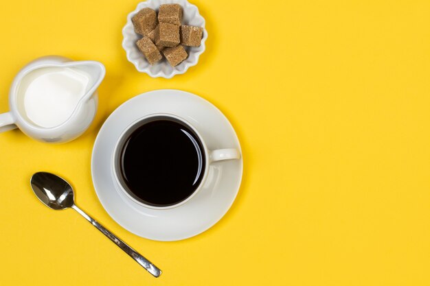 A cup of coffee with a milkmaid and brown sugar cubes on a yellow background