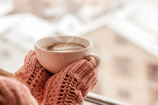 冬の雪に覆われたウィンドウの背景にニットのセーターに女性の手で蒸気とコーヒーのカップ