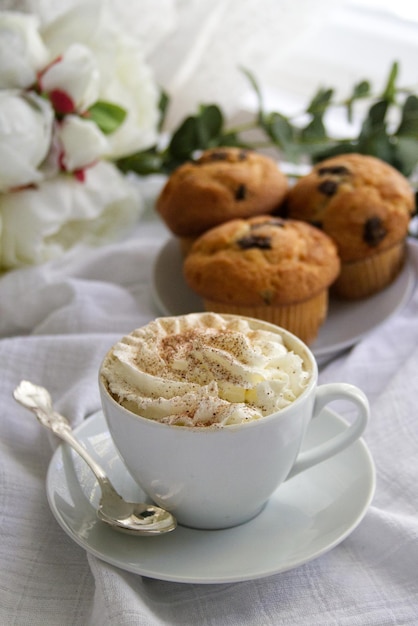 A cup of coffee with whipped cream next to a plate on which there are muffins with chocolate