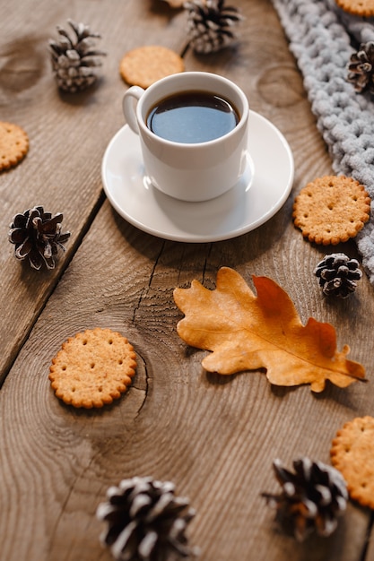 A cup of coffee on a wooden table with cookies, leaves and cones