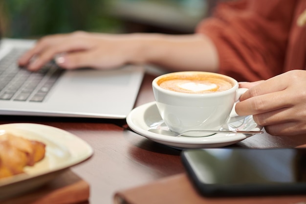 Cup of delicious cappuccino on table of female freelancer working from home