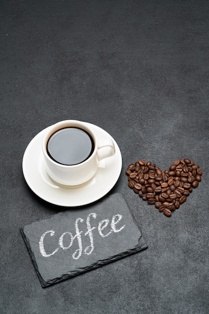 Cup of espresso and heart shaped roasted coffee beans on dark concrete table