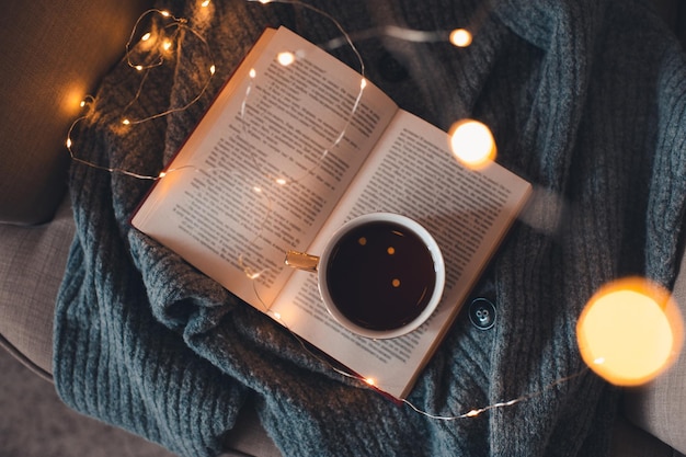 Tazza di tè nero fresco soggiorno su libro di carta aperto con maglione di lana a maglia in una sedia accogliente sopra il bagliore delle luci di natale primo piano. vista dall'alto. colazione mattutina. ambiente familiare invernale confortevole.