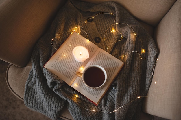 Cup of fresh black tea stay on open paper book with knit woolen sweater in cozy chair over glow Christmas lights closeup. Top view. Early morning breakfast. Winter comfy home atmosphere.