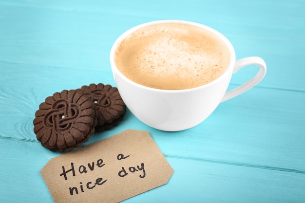 Cup of fresh coffee with cookies and card on light blue background