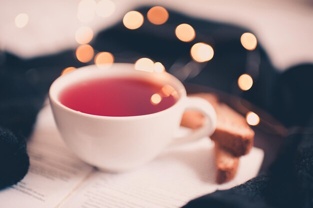 Cup of fresh herbal tea on open book with tasty cookies over Christmas lights at background. Good morning. Merry Christmas and Happy New Year.