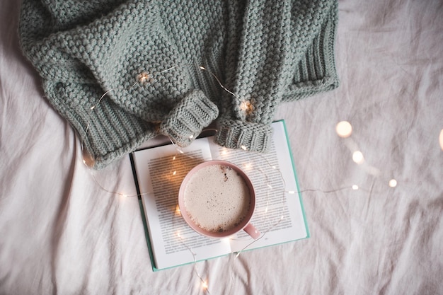 Cup of fresh hot coffee staying on open book with knitted sweater in bed closeup. Top view. Winter season.