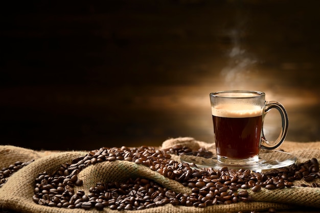 Cup glass of coffee with smoke and coffee beans on burlap sack on old wooden table