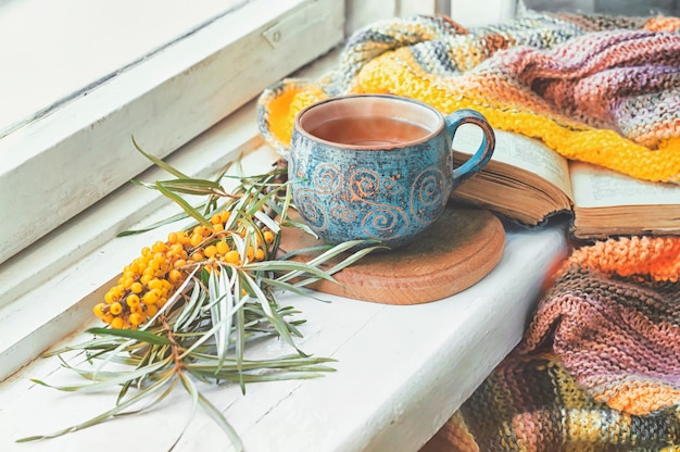 Чашка травяного чая, облепиха и вязаное одеяло на подоконнике.