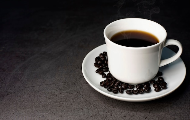 A cup of hot black coffee on a black background with roasted coffee beans and hot steam coming out of the mug.