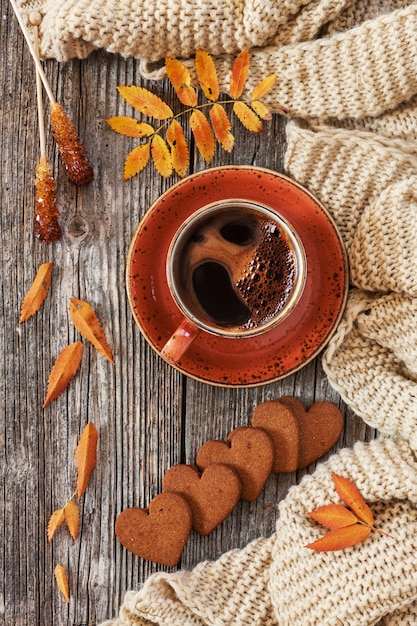 Cup of hot black coffee and heart-shaped gingerbread with autumnal leaves and warm scarf. 