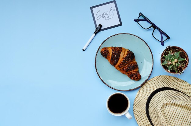 Cup of hot black coffee, notebook, sweet croissant, cacti close up on blue background. top view