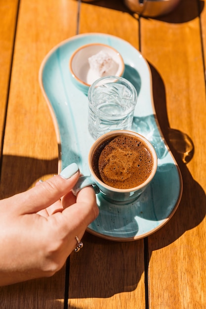 A cup of hot black turkish coffee in blue cup in women's hands served with a glass of water and sugar