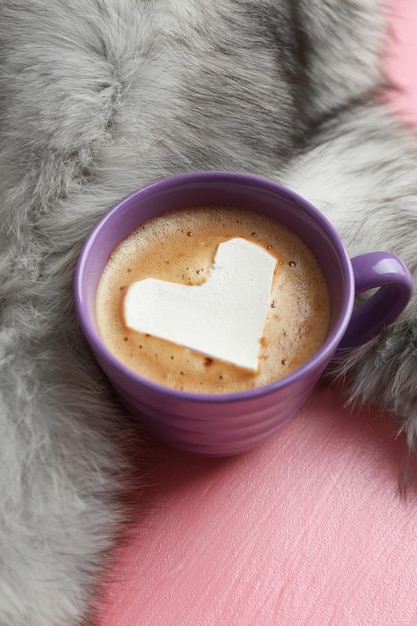 Cup of hot cappuccino with heart marshmallow and fur on pink background close up