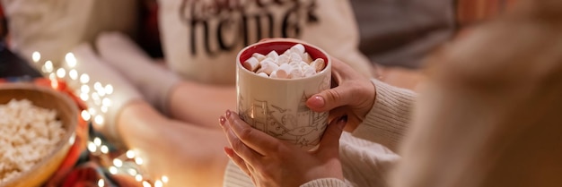 Cup of hot chocolate or coffee drink with marshmallows in female holding hands of woman in knitted sweater relaxing and enjoyment the holidays in bed home on christmas or new year holidays eve banner