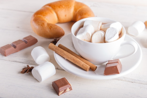 A cup of hot chocolate with marshmallow, croissant and spices on white wooden background.