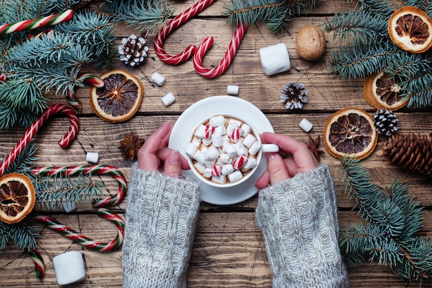 A Cup of hot chocolate with marshmallow in the hands of a man in a sweater. Christmas tree and decorations, cane caramel and oranges nuts Wooden background copy space. New year gift.