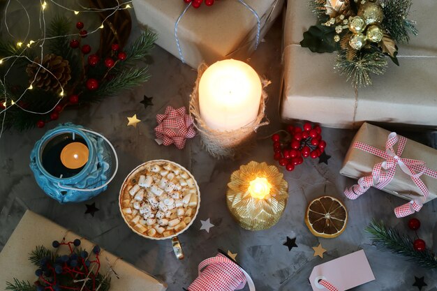 A cup of hot cocoa with marshmallow among Christmas gifts next to candles 