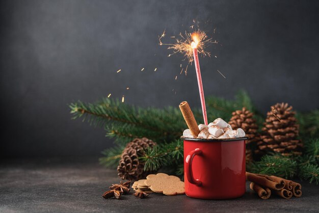 Cup of hot cocoa with marshmallow and cinnamon stick surrounded by spruce branches