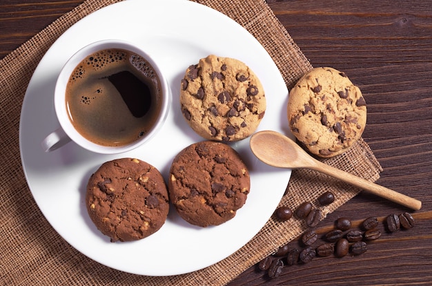 Cup of hot coffee and cookies with chocolate and nuts in plate on brown wooden table, top view