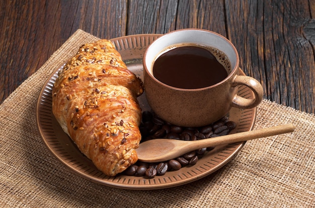 Cup of hot coffee and croissant with sesame seeds in plate on dark wooden table