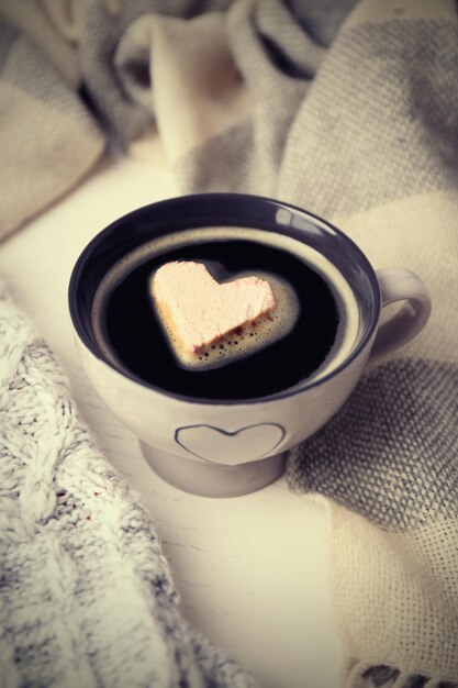 Cup of hot coffee with heart marshmallow and warm scarfs on light wooden background close up