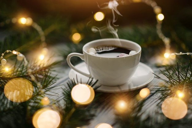 A cup of hot coffee with marshmallows among the fir branches and lights of festive garlands. Winter composition with selective focus.