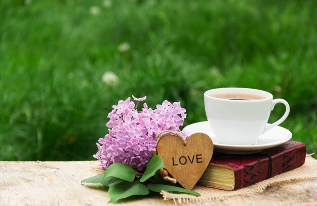 Cup of hot tea, book and lilacs against background of green grass