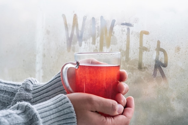 A cup of hot tea in the hands of an adult by the window on a cold winter day