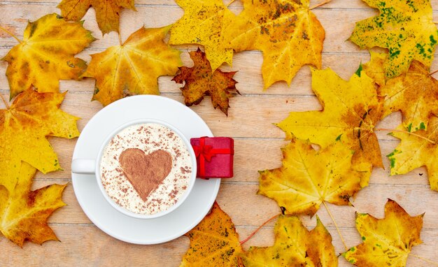 Фото Чашка кофе с сердцем из корицы и подарочной коробкой на столе, вокруг кленовые листья