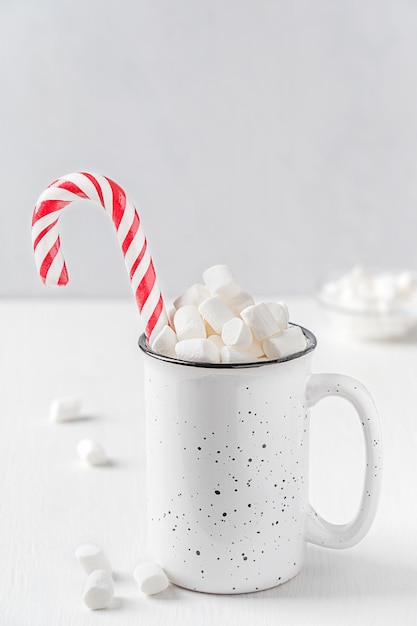 Фото Чашка домашнего сладкого горячего какао-напитка с зефиром и полосатой конфетной тростью на белом столе