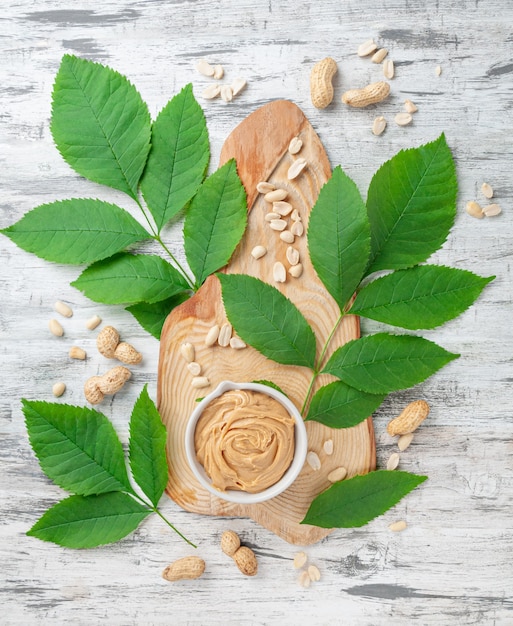 A cup of peanut butter On a wooden textured board. Decorated with greens and peanuts.