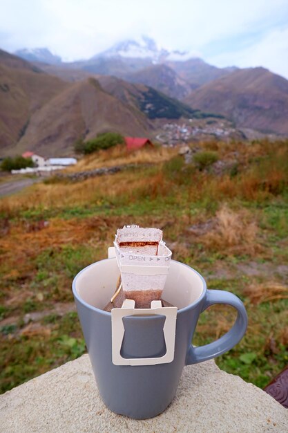 背景にぼんやりした村の景色が映っている屋外のテラスで準備されているポータブルドリップコーヒーのカップ