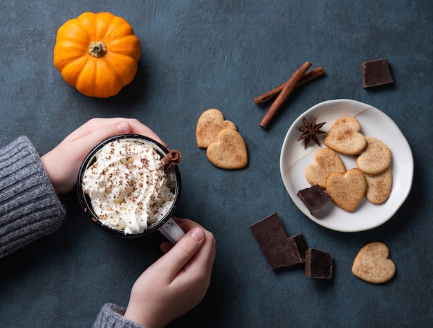 Una tazza di latte di zucca con crema in mano di donna su un tavolo scuro con biscotti, cioccolato e cannella