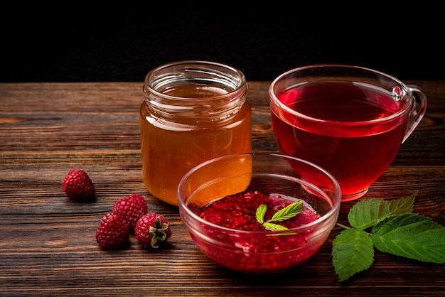 Cup of raspberry tea with raspberry jam and honey on dark wooden background.