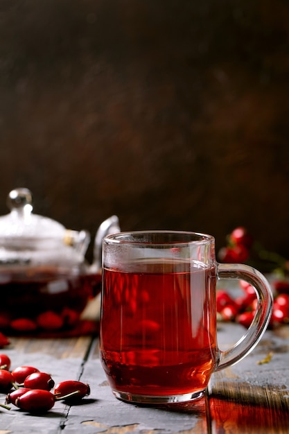 Tazza di tisana di bacche di rosa canina e teiera di vetro in piedi su un vecchio tavolo di legno con bacche autunnali selvatiche intorno. bevanda calda vitaminica.