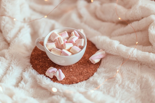 Cup of tasty cocoa drink or hot chocolate with marshmallows in cup on cork stand on a white bedspread. Winter dessert beverage. Decoration garlands of lights.