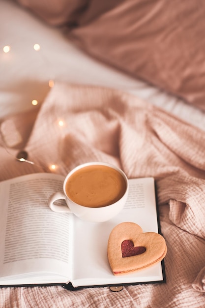 Cup of tasty coffee with red heart shape cookie on open paper book in bed over Christmas lights close up. Good morning. Breakfast. Valentines Day.