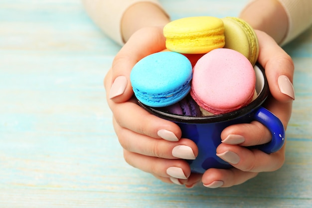 Cup of tasty colorful macaroons in female hands on color wooden background