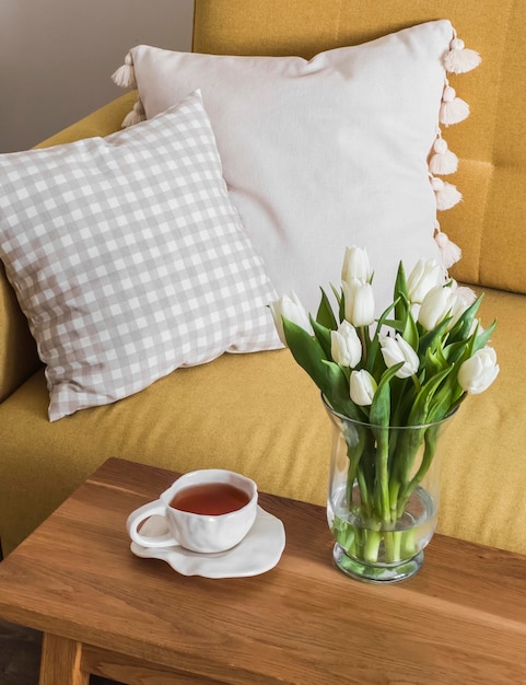 茶のカップソファの近くの木製のベンチにあるガラスの花瓶の中のチューリップの花束