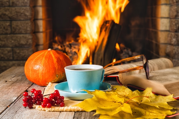 A cup of tea or coffee, pumpkin, autumn leaves and viburnum berries before cozy fireplace, in country house, autumn or winter holidays.