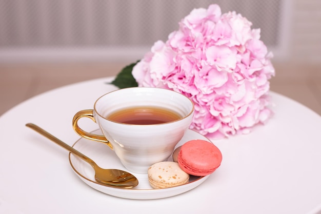 Cup of tea, macaroons, golden knife and fork and pink hydrangea flower on white table