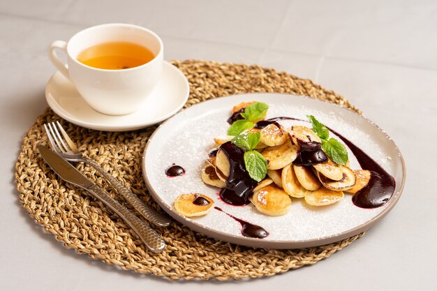 A cup of tea and mini-pancakes with berry jam and mint sprinkled with powdered sugar.