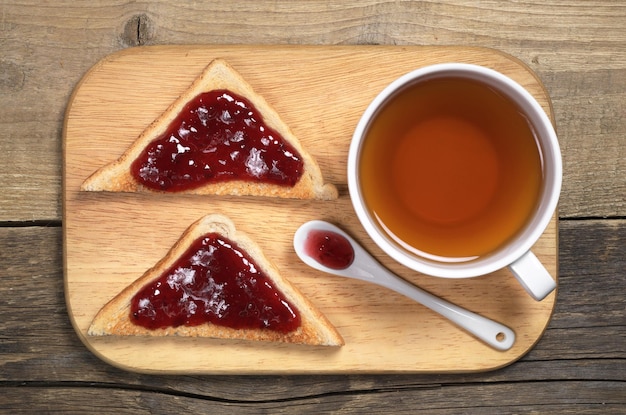 오래된 나무 탁자 위에 있는 커팅 보드에 딸기 잼을 얹은 차 한 잔과 구운 빵