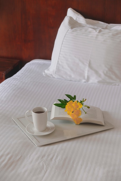 Cup of tea with a book on a white tray with a yellow flower on a white bed