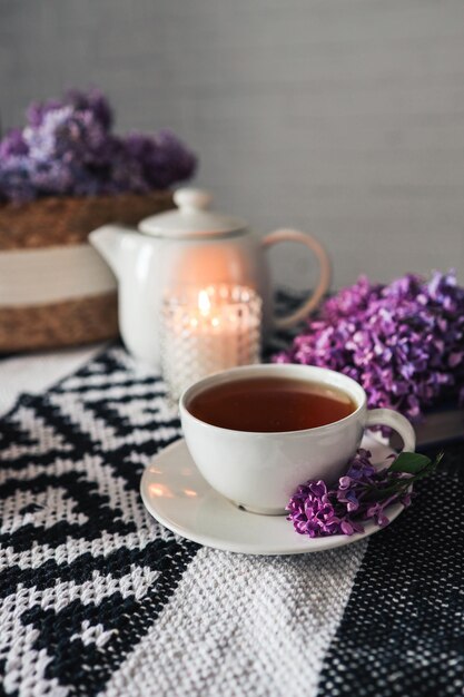 A cup of tea with a branch of lilac on a saucer on the table Candle teapot wicker basket