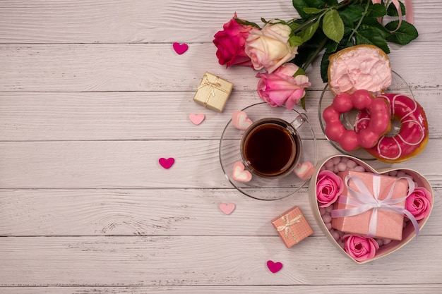 Cup of tea with donuts, fresh roses, more gifts, red ribbon on wooden table for Valentines Day