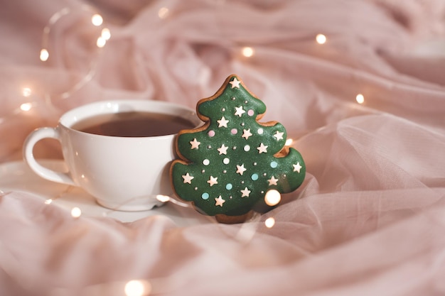 Cup of tea with gingerbread in Christmas tree shape closeup over glowing lights. Winter holiday season. Xmas.